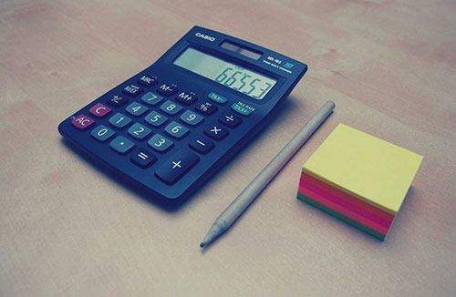 A handheld calculator, pen, and pad of paper on table top.