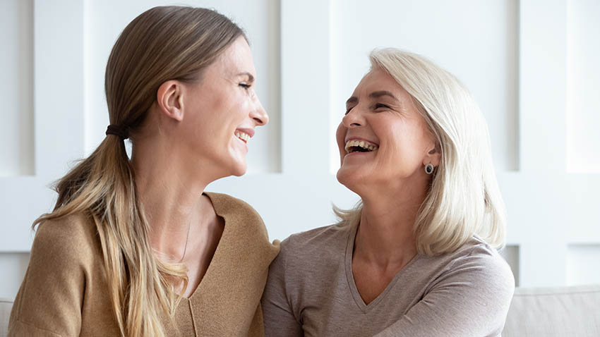 Happy older mom and grownup daughter sitting on couch talking and laughing at home